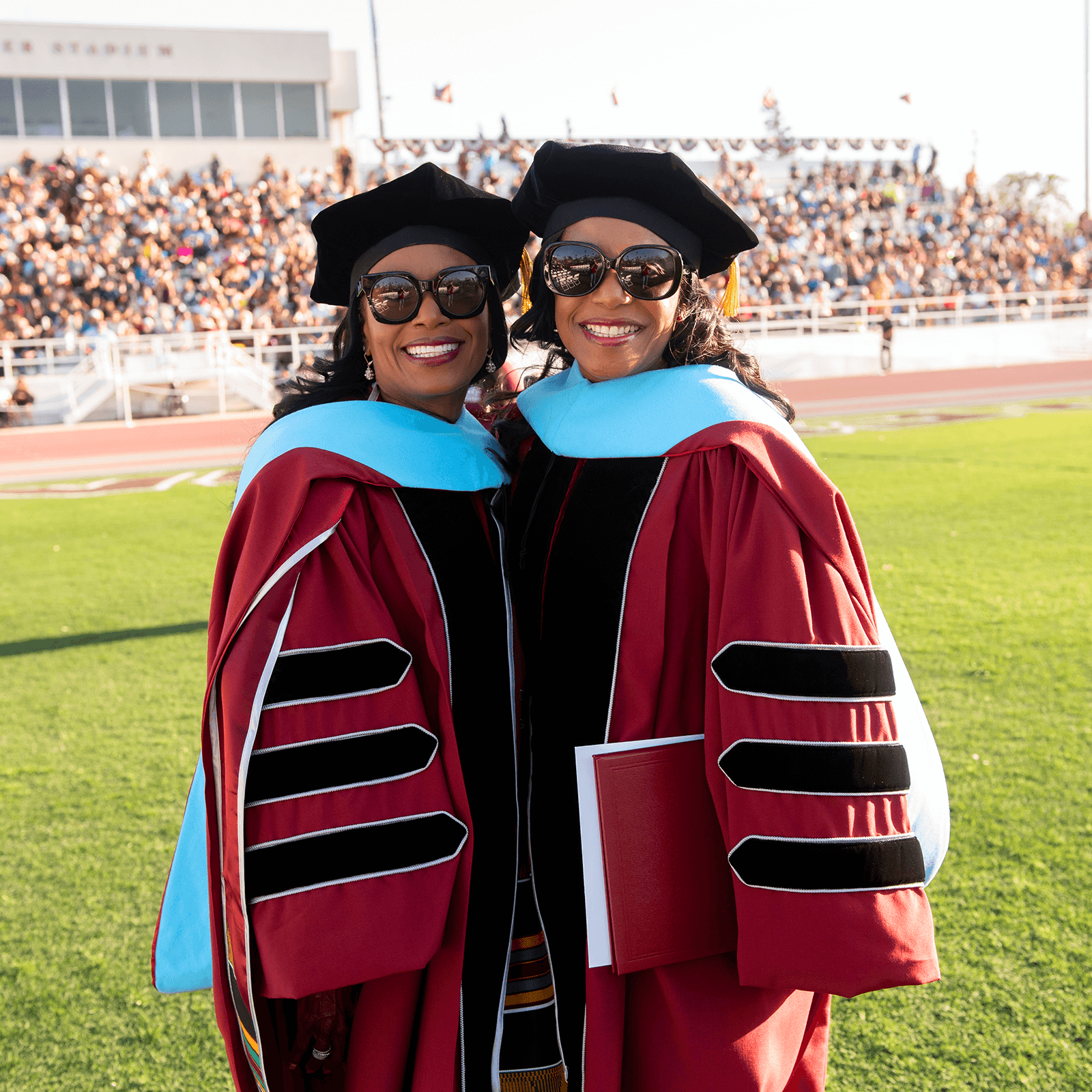 two women in graduation gowns and cap and gowns standing on a field with a crowd of people