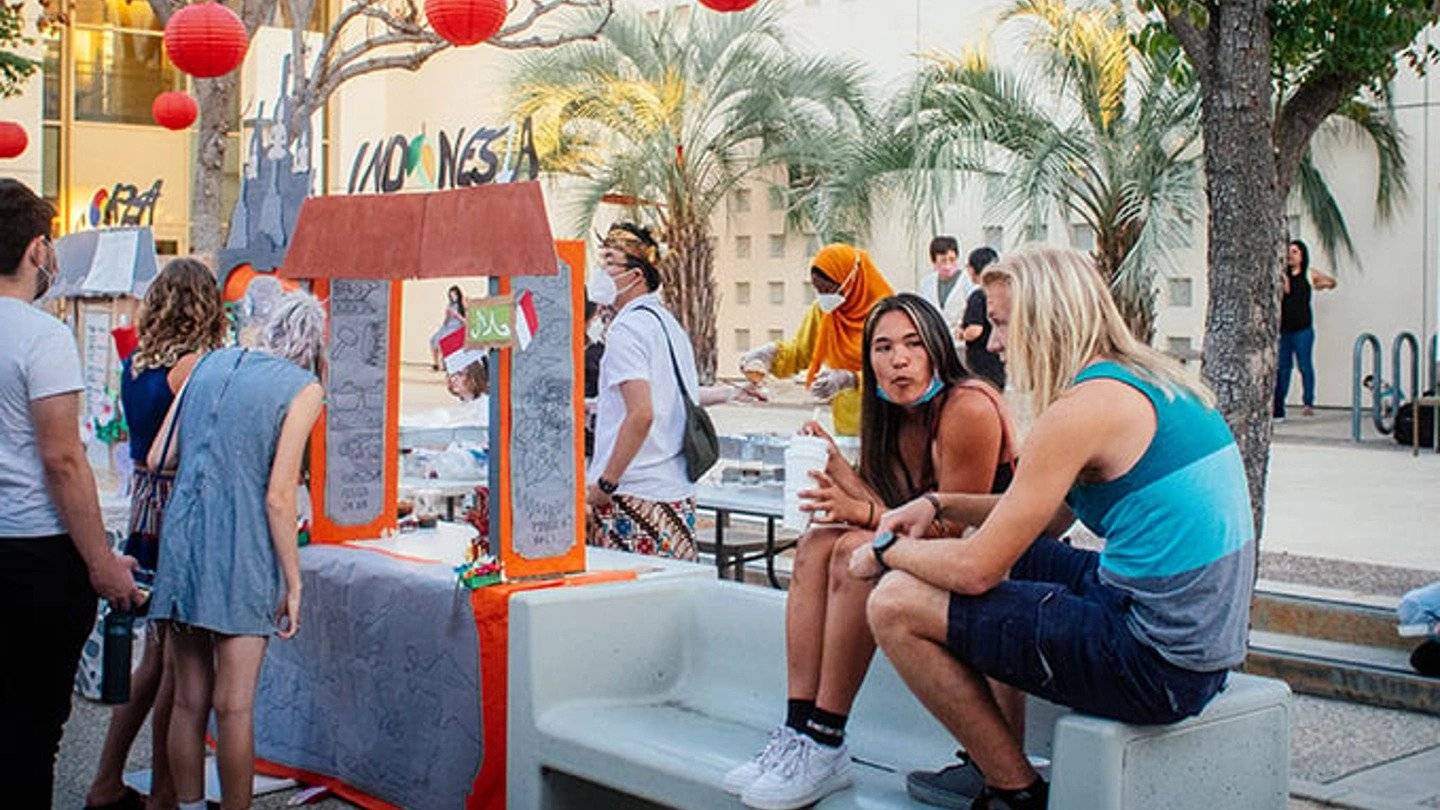 A group of people walking around at the night market, while two attendees sit on a bench.