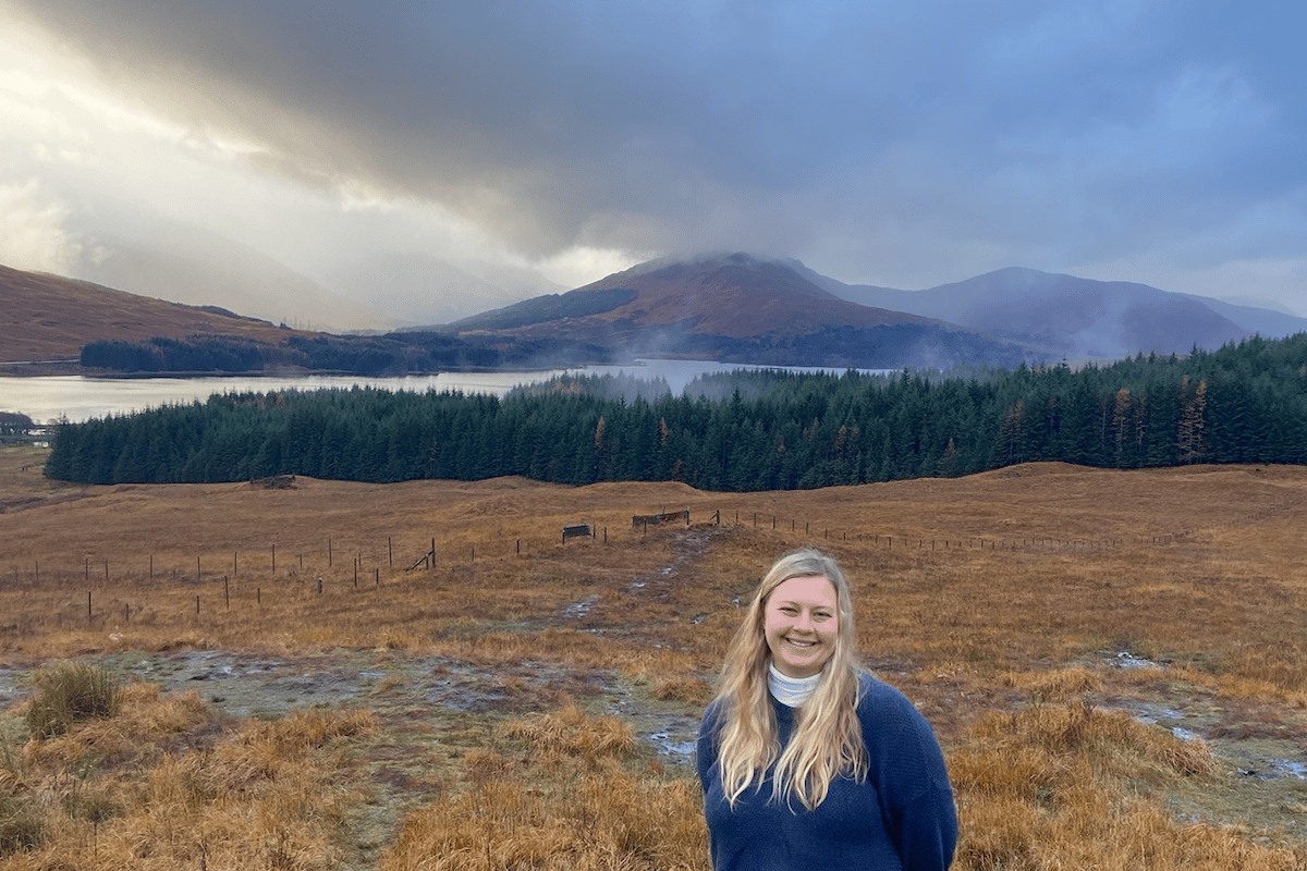 a woman standing in a field with trees and mountains in the background