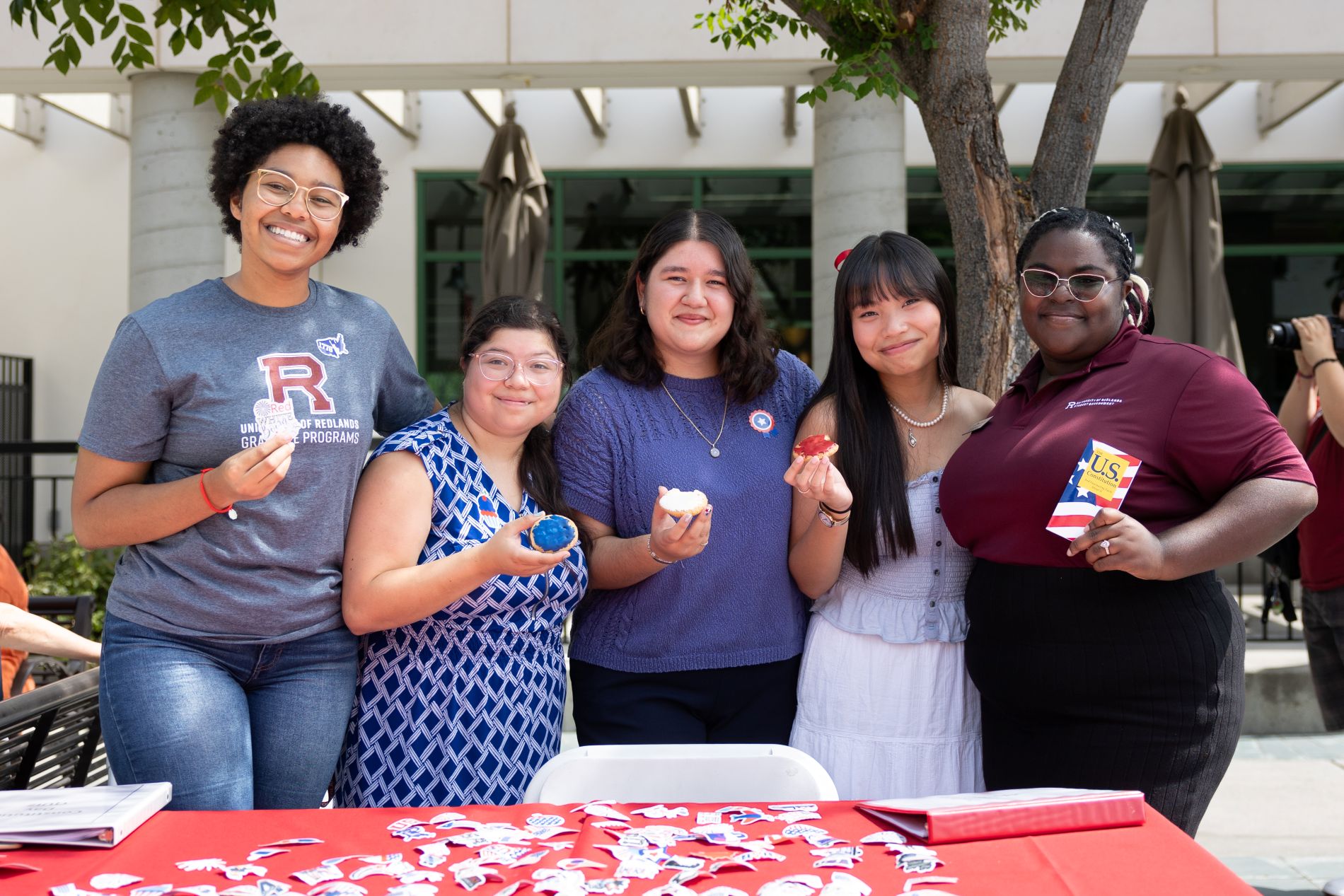 Five students stand together behind a table with stickers and cupcakes at a campus gathering.