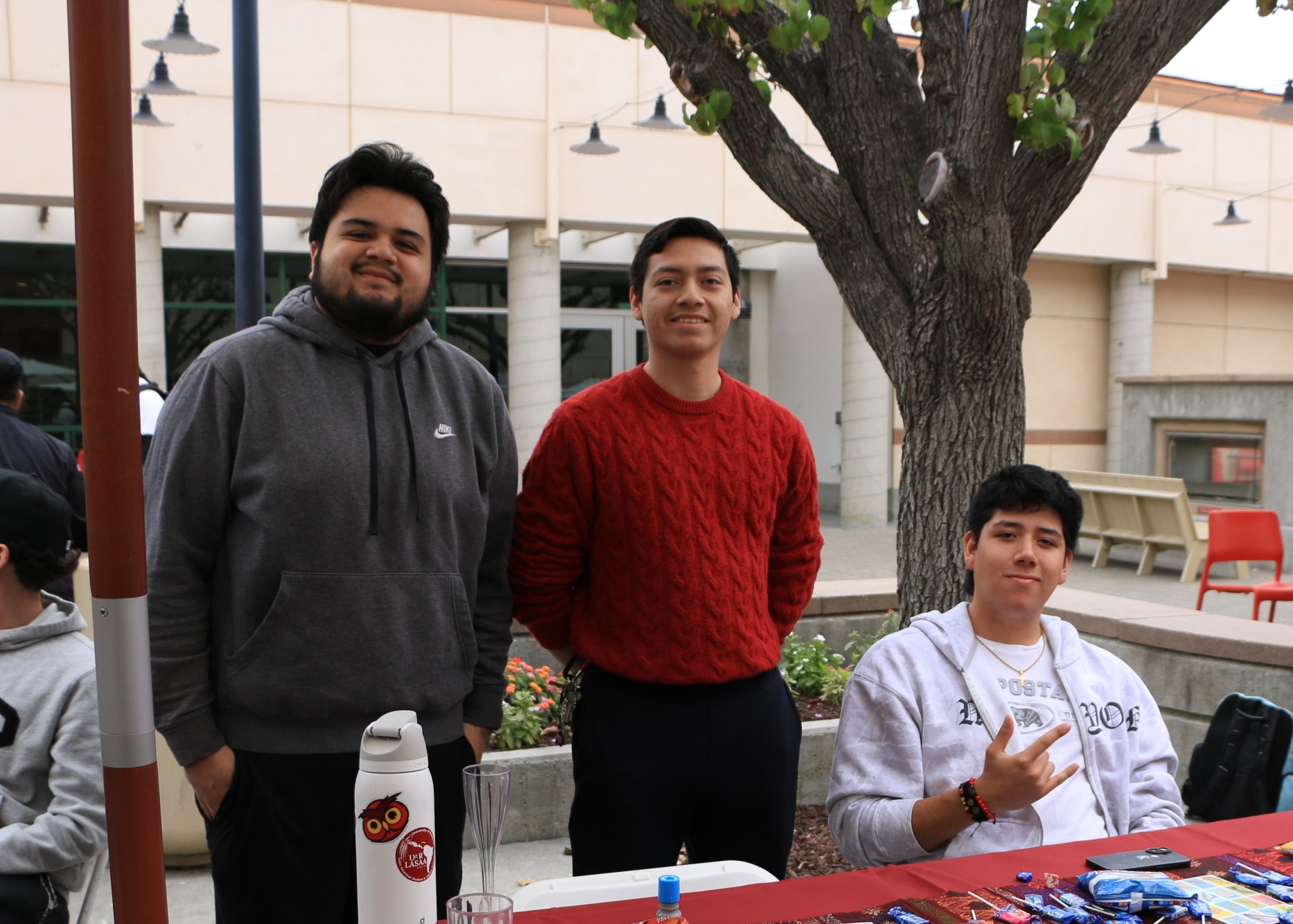 Three students pose behind a campus organization booth with informational materials and snack giveaways on the table.
